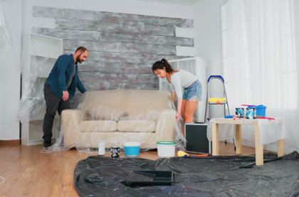 A husband and wife wrapping a piece of furniture in protective wrapping, with paint, a ladder and plastic tarp in the background