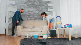 A husband and wife wrapping a piece of furniture in protective wrapping, with paint, a ladder and plastic tarp in the background